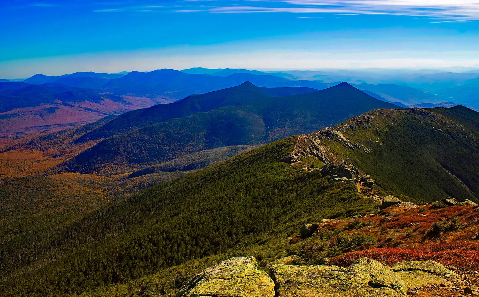 A colored photograph of a mountain region. The mountains all have trees with leaves colored in green, red, yellow, and orange.