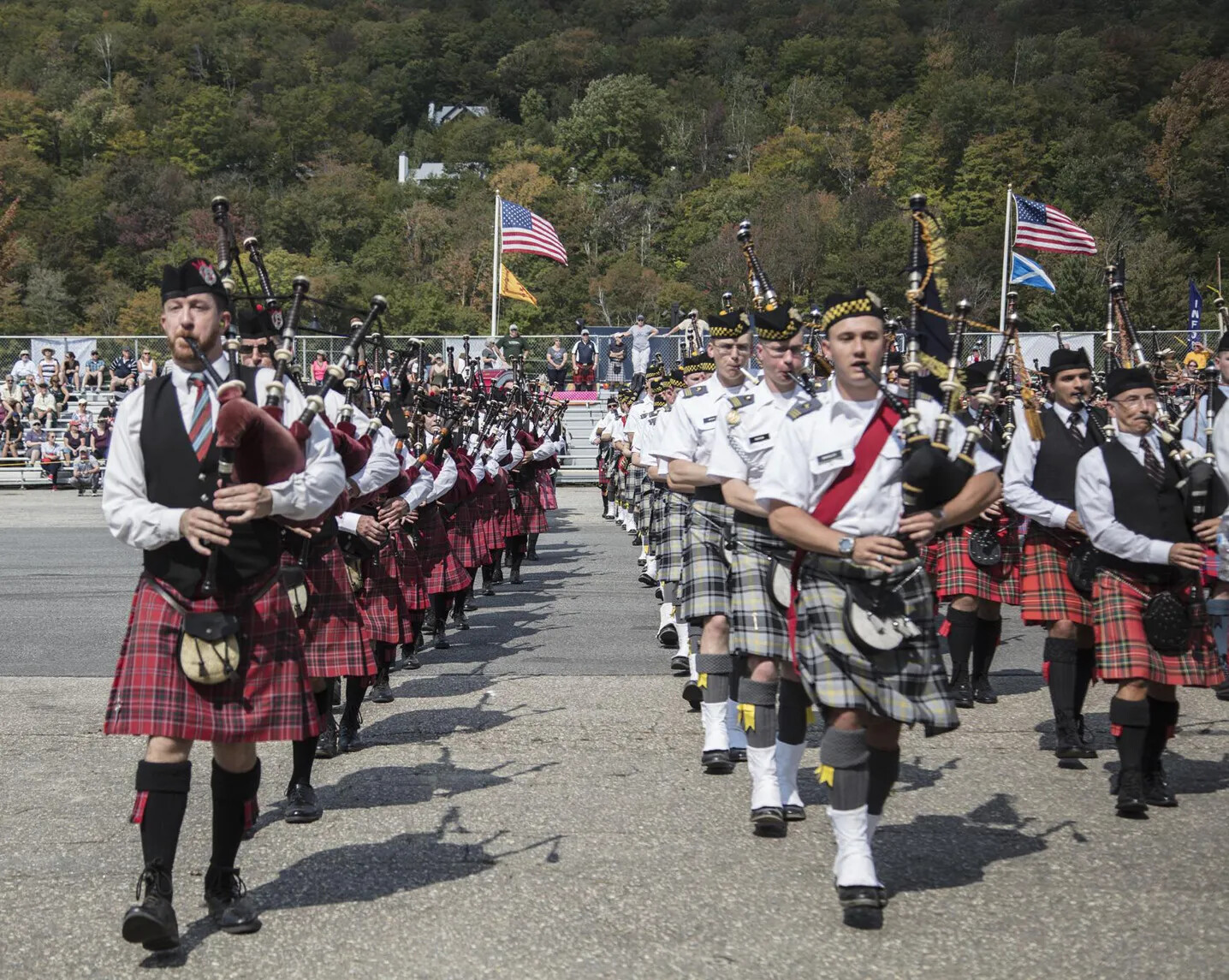 A colored photograph of a group of white men in white shirts and different colored kilts playing bagpipes. They all stand in three straight lines.