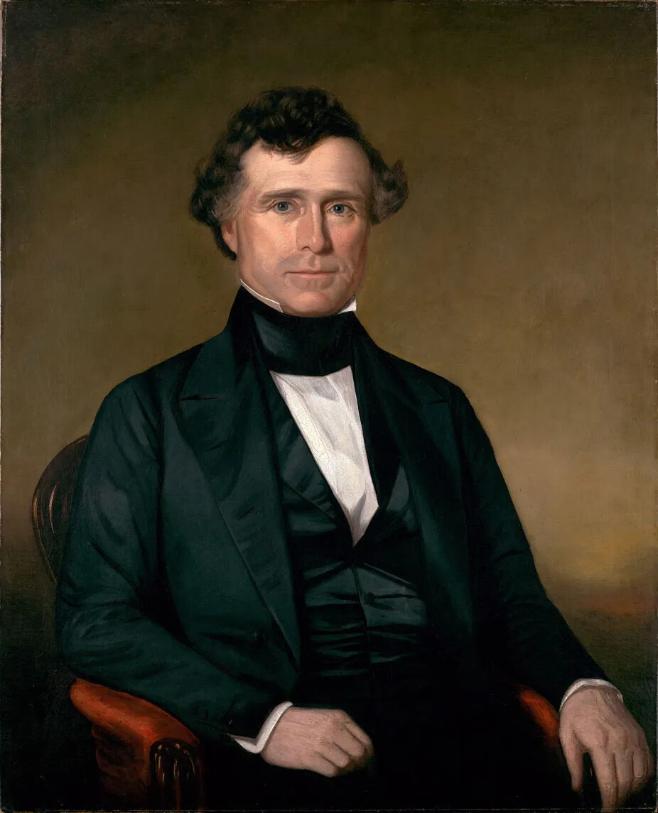 In a color painting, a man sits on a leather chair against a brown background. He is wearing a black suit in the mid-1800s style with high collar and no tie. His hands rest on the arms of the chair in a casual manner, and he looks directly at the viewer with a small smile. He has short curly brown hair and no facial hair. 