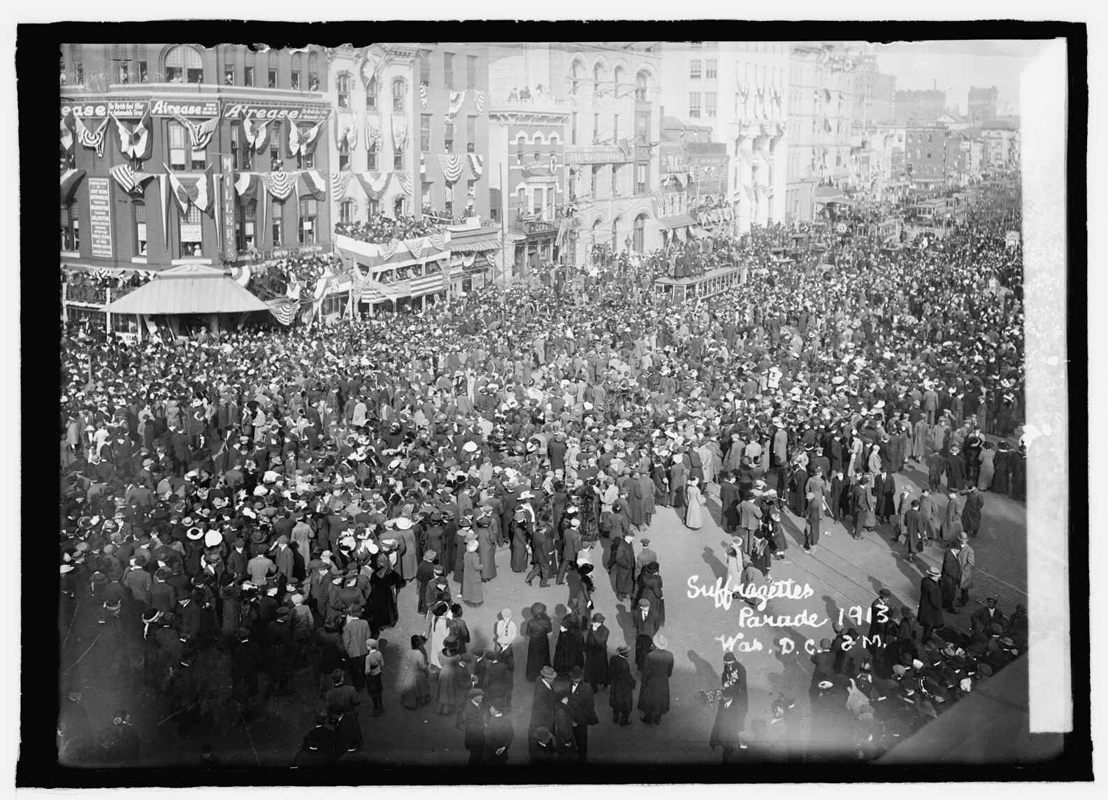 A black and white photograph shows a broad city street filled with people and no cars, seen from above. People generally wear long skirts or coats and hats. The buildings are decorated with half-circle flags draped from windows. Words in cursive in the lower right hand corner read "Suffragettes Parade 1913 Was. D.C. [illegible]." 