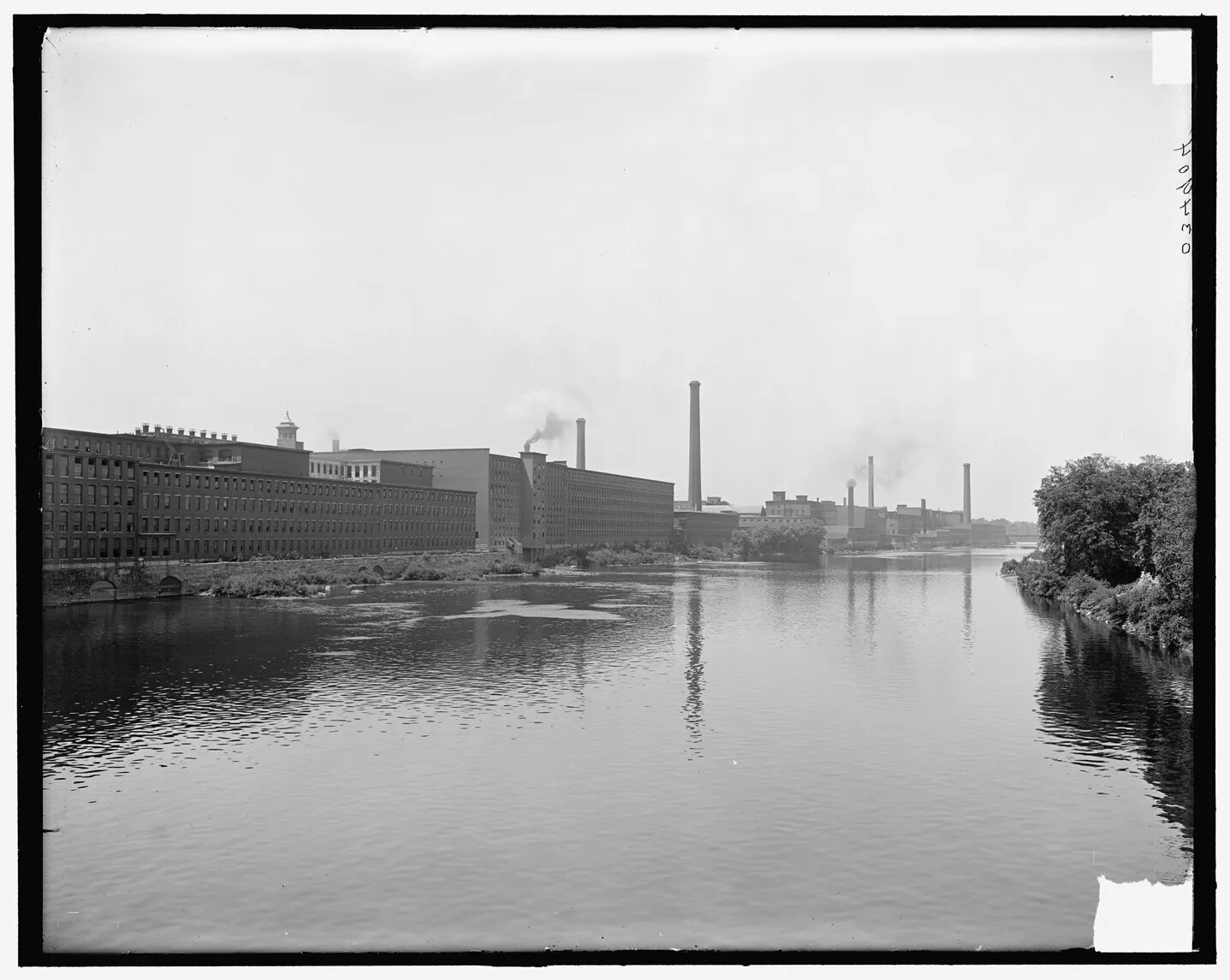 A black and white photograph shows a wide, calm river in its center. On the right, the riverbank is lined with trees. On the left, long industrial buildings about five stories tall run the length of the river. There are many big windows on the buildings and they are flat on top. Smoke stacks are seen in the distance. 