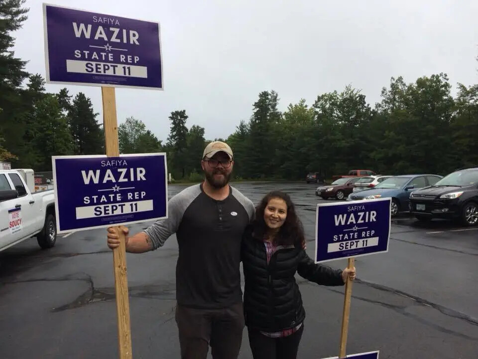 Two people stand in a parking lot in a color photograph. A woman wears a black jacket and smiles, and a smiling man wears a cap, and casual pants and shirt. He holds two signs that are the same stacked on each other on a wooden pole, and she holds one of the same sign. The signs read “SAFIYA/ WAZIR/ STATE REP/ SEPT 11.” 