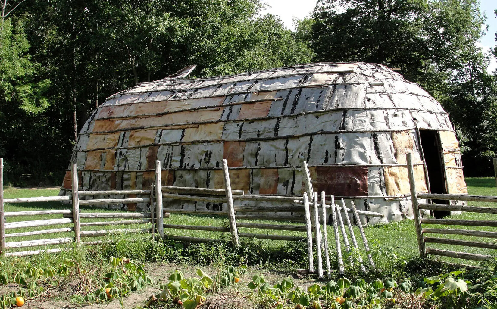 A long enclosure made from wood and animal skins, placed on a grassy field. In front of the enclosure is a garden fenced in by wooden posts.
