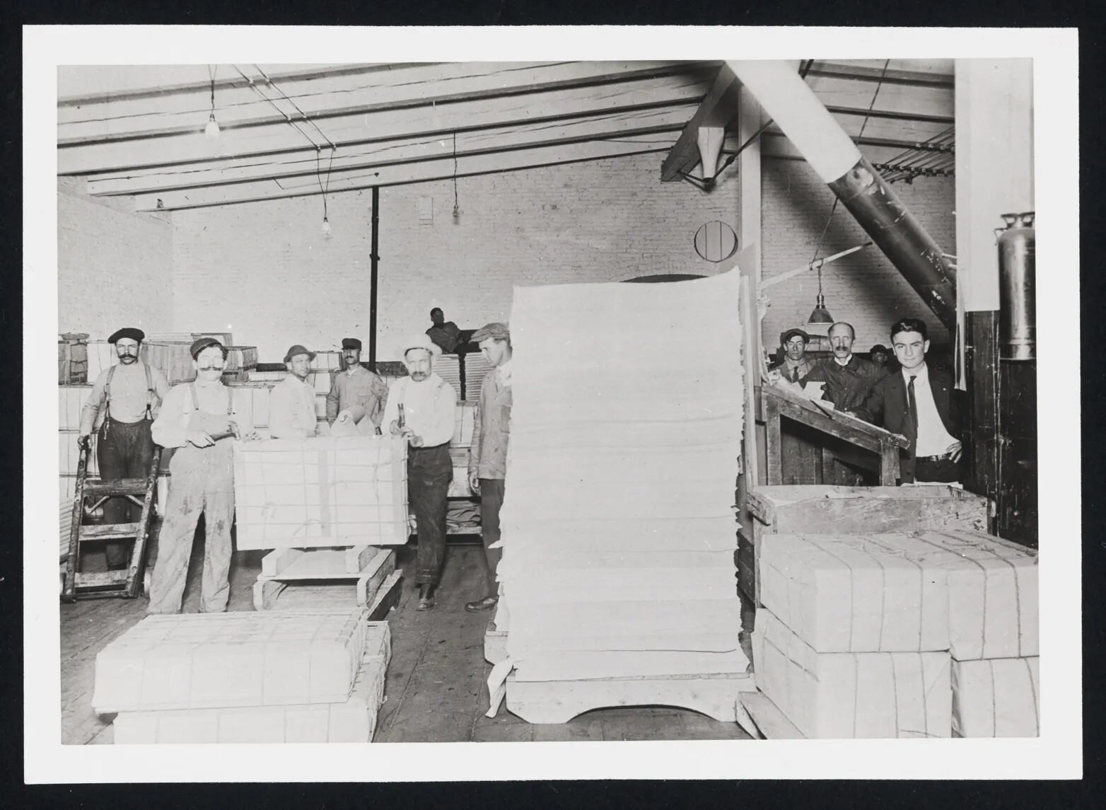 A black and white photograph shows the inside of a small factory. Men in caps and working clothes are shown gathered around stacks of white paper of various heights. The stacks are about three feet wide, and one of the stacks is taller than a person.   