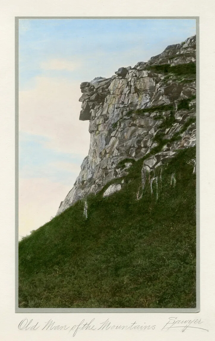 A colorized photograph of a rocky cliff. At the cliff's edge, the rocks come together to form the profile of a man's face. Green trees are seen at the bottom of the cliff. 
