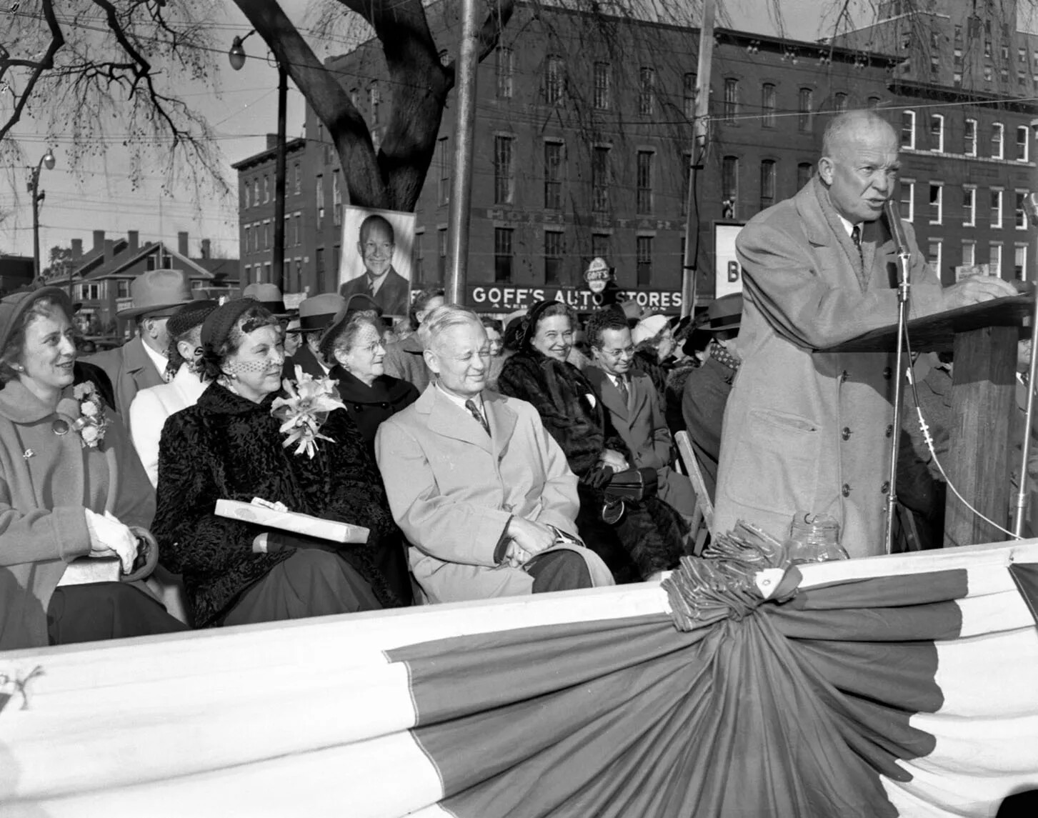 A black and white photograph of a man standing a microphone to the right and a group of people seated behind him. Someone holds up a large poster with a picture of the man speaking on it. Behind the group are tall brick buildings and houses.