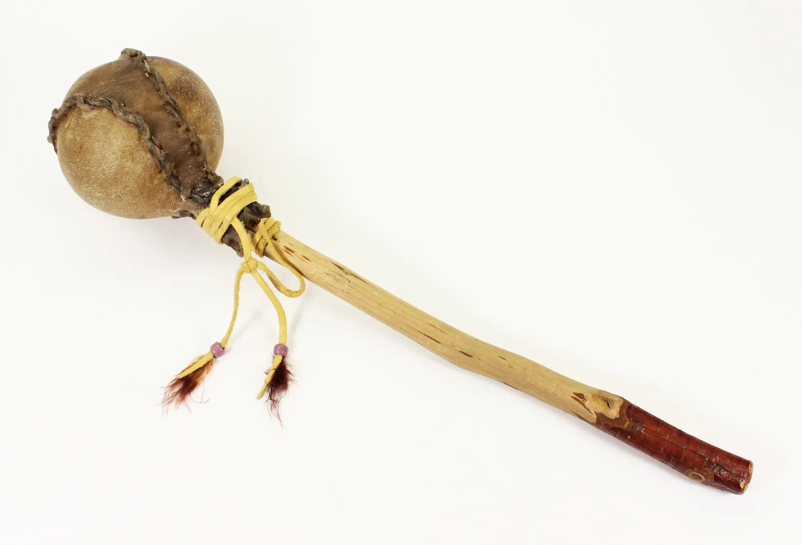 A large brown rattle with a long wooden handle. In between the rattle ball and handle is a yellow piece of rope tied around the handle, with purple beads and feathers attached to the ends of the rope.